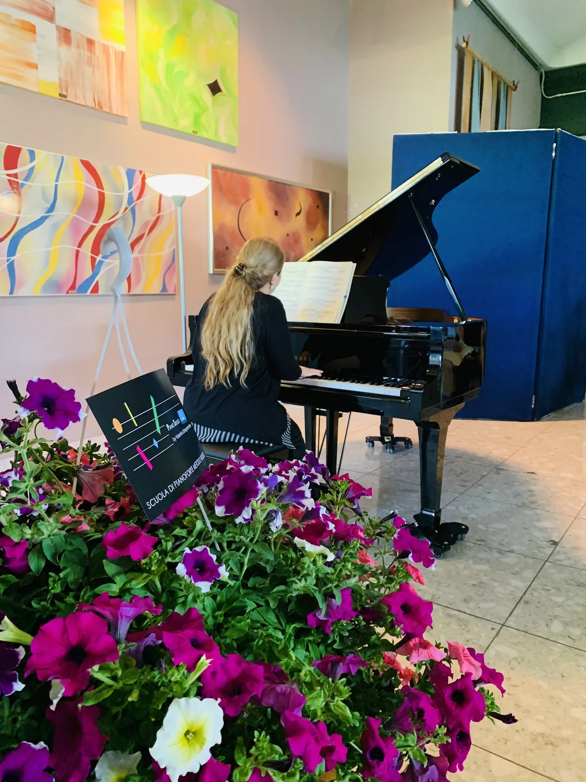 Un'allieva suona al pianoforte a coda, di spalle, dietro un vaso di petunie